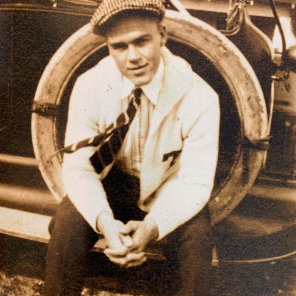 A sepia-toned photograph of a white male in early adulthood seated outdoors. He is wearing a white letter sweater with a T on the breast.