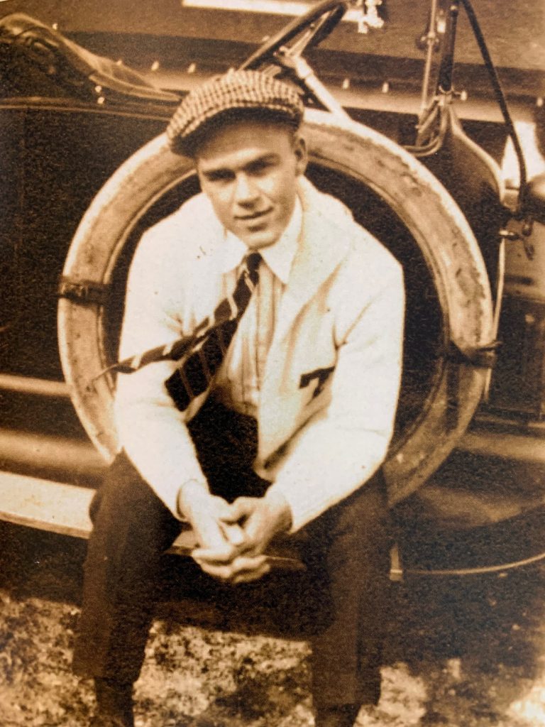 A sepia-toned photograph of a white male in early adulthood seated outdoors. He is wearing a white letter sweater with a T on the breast.
