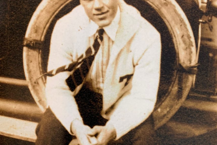 A sepia-toned photograph of a white male in early adulthood seated outdoors. He is wearing a white letter sweater with a T on the breast.