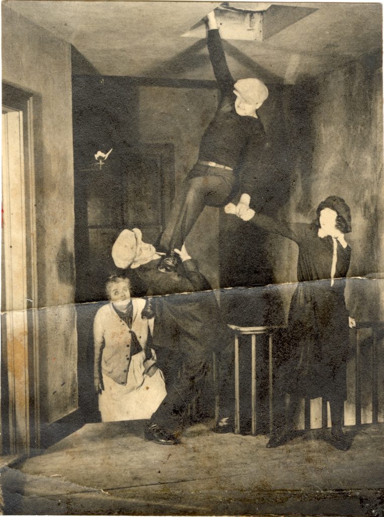 Joe Bonomo hanging onto ceiling, holding young woman's hand and pushing down on another man with his feet. Older woman looks on.