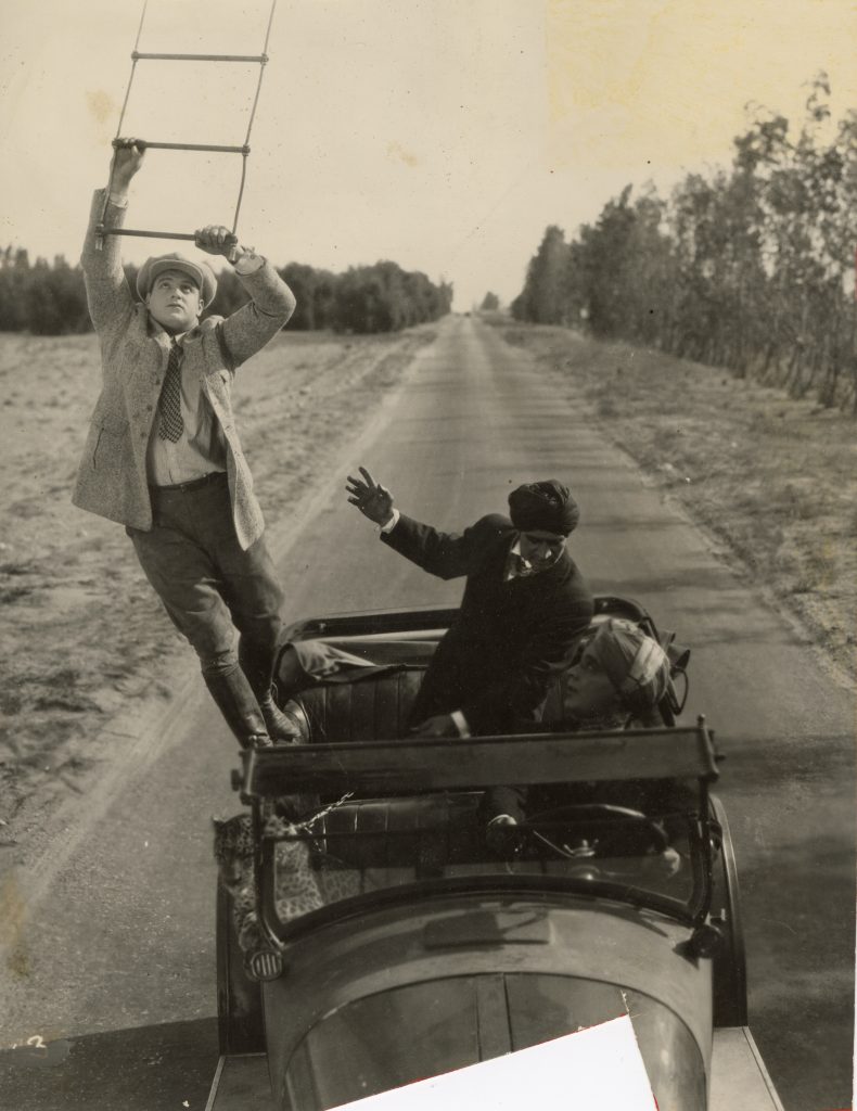Joe Bonomo climbing onto ladder from a moving vehicle. Two men and a cheetah are in the vehicle. 