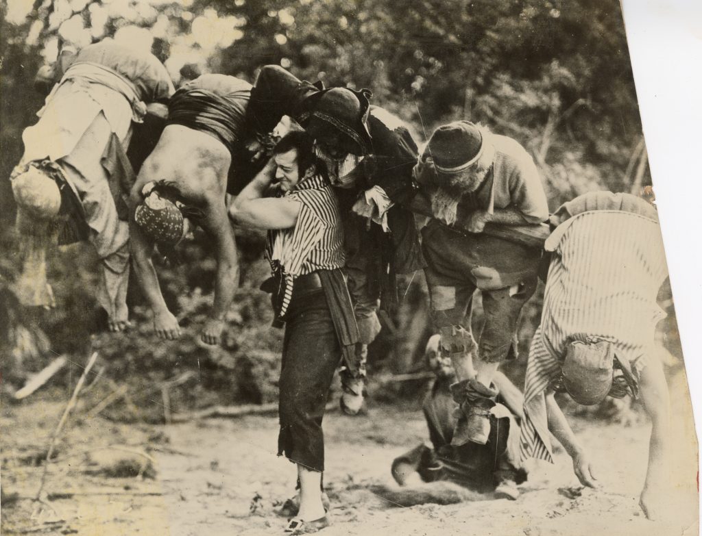 Joe Bonomo carrying log that has 5 men hanging onto it. One man seated looks on.