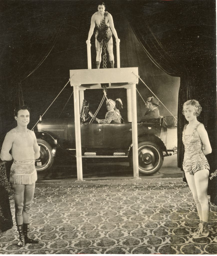 Joe Bonomo standing above platform with chains around his waist. Chain attached to ropes holding a car below with 4 seated men. A man and a woman stand in front looking at the camera. 