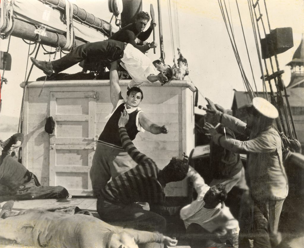 Joe Bonomo standing on boat lifting one man over his head with one arm. Other man surrounding him. 