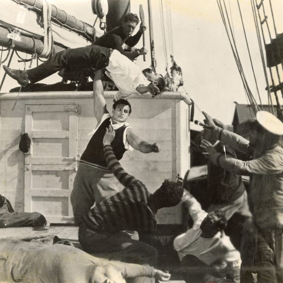 Joe Bonomo on a boat surrounded by men, lifting one man over his head with one arm.