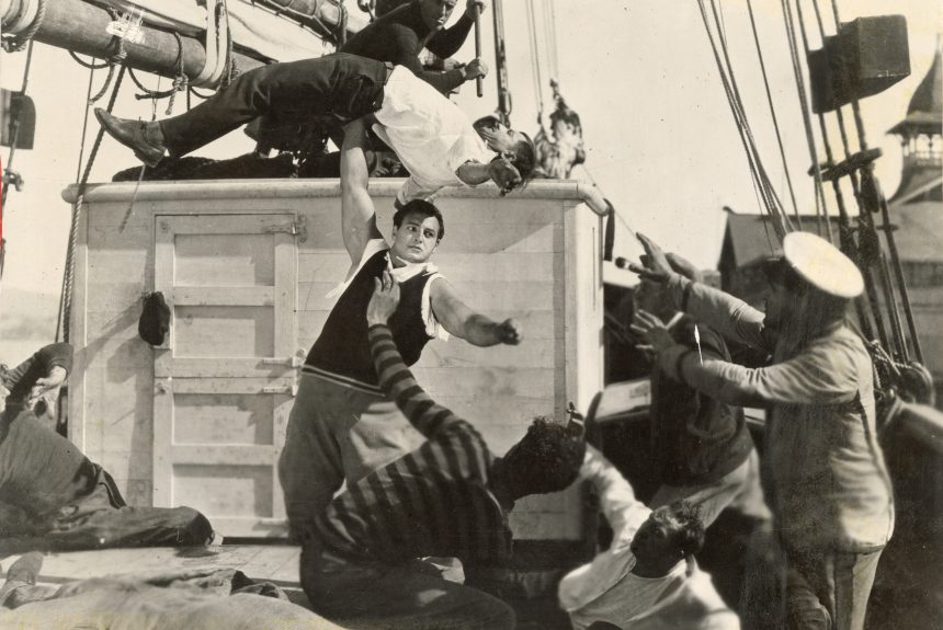 Joe Bonomo on a boat surrounded by men, lifting one man over his head with one arm.