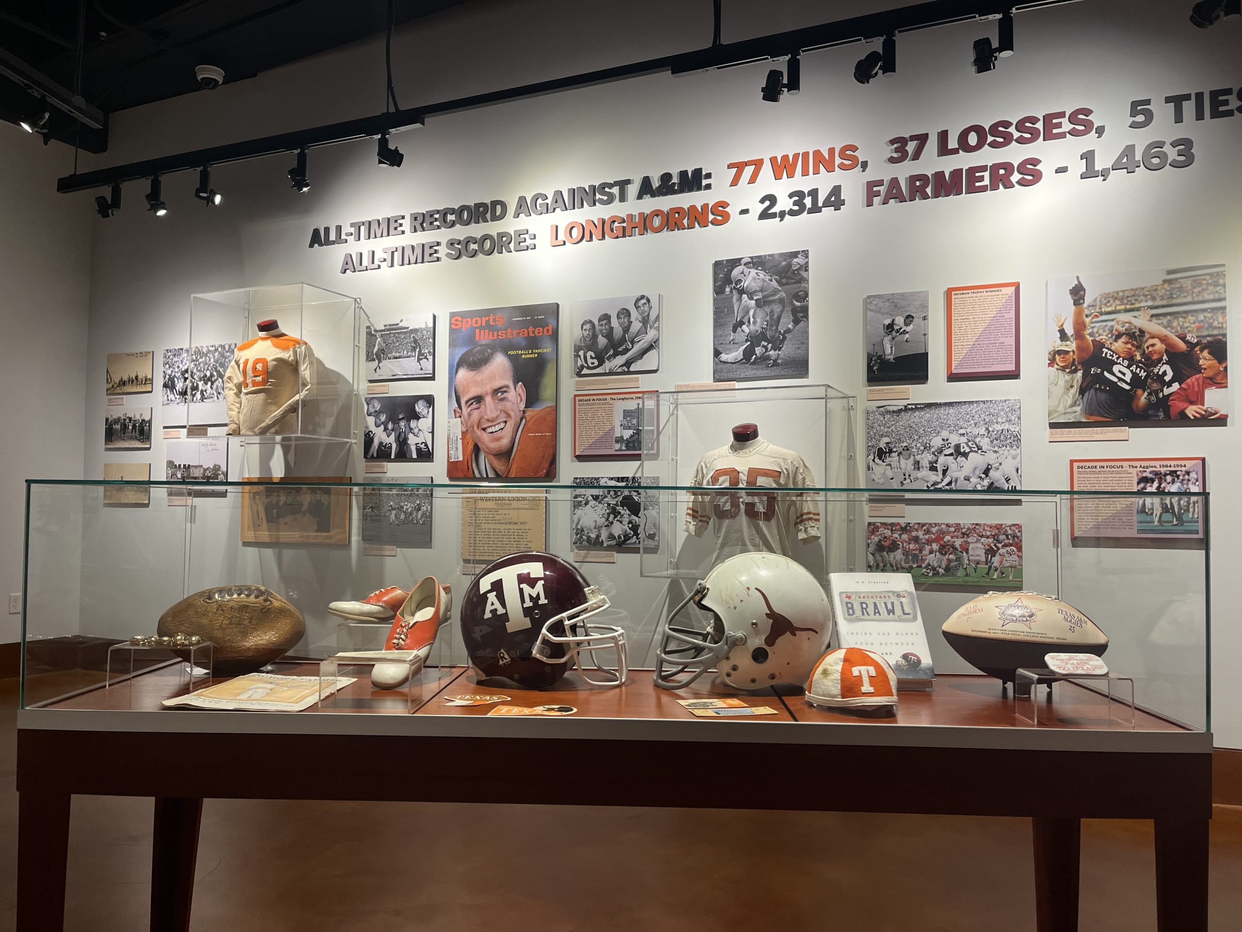 A photo of the table case in the center of the gallery, with Aggie and Longhorn helmets facing each other, as well as historic footballs, and other memorabilia.