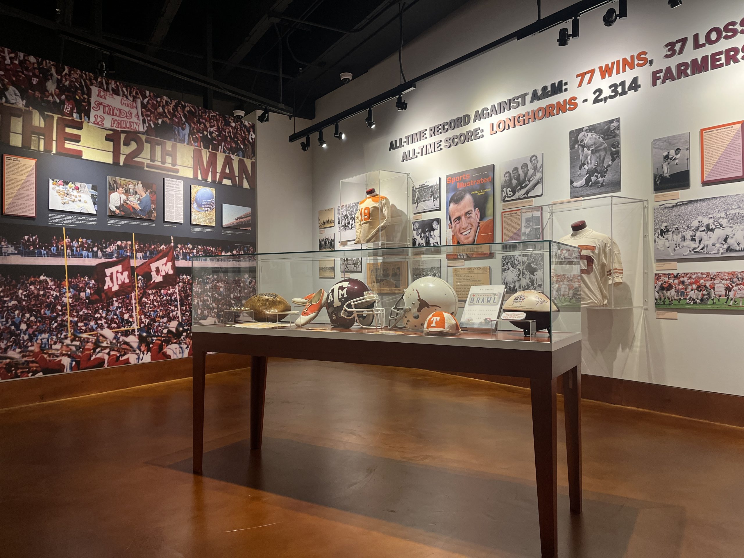 A color photo of the new exhibit in the gallery, a table case in the foreground with artifacts and football helmets, photos and 3D lettering on the walls in background.
