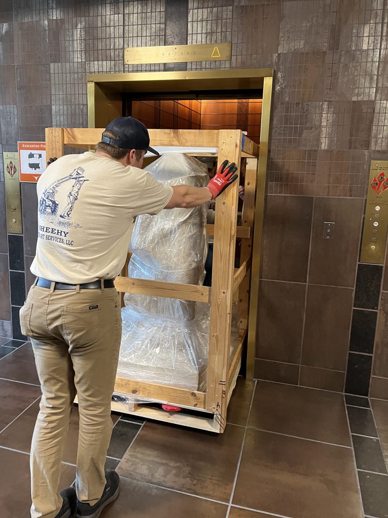 A man helps push a crated sculpture on a pallet jack into an elevator for delivery.