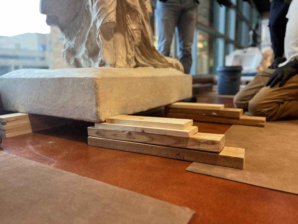 Step two of lowering the base of Winged Victory to the ground. It now sits on wood boards, the pallet jack has been removed.