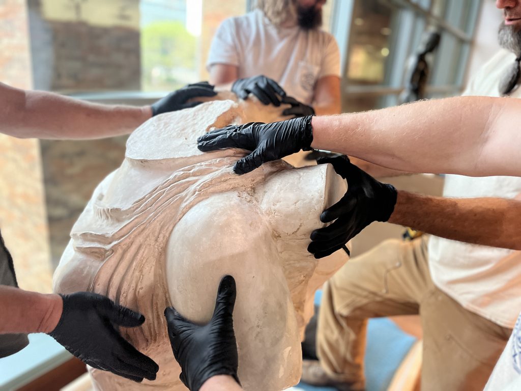 The many black-gloved hands of the Sheehy Fine Art Services crew all placed on the top half of the Winged Victory sculpture as they prepare for the next step in assembly.