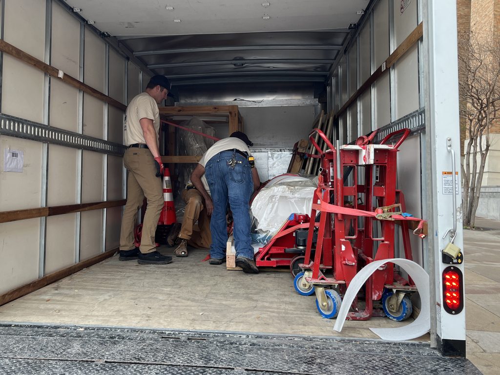 Three men unload a crated sculpture from the back of a box truck.