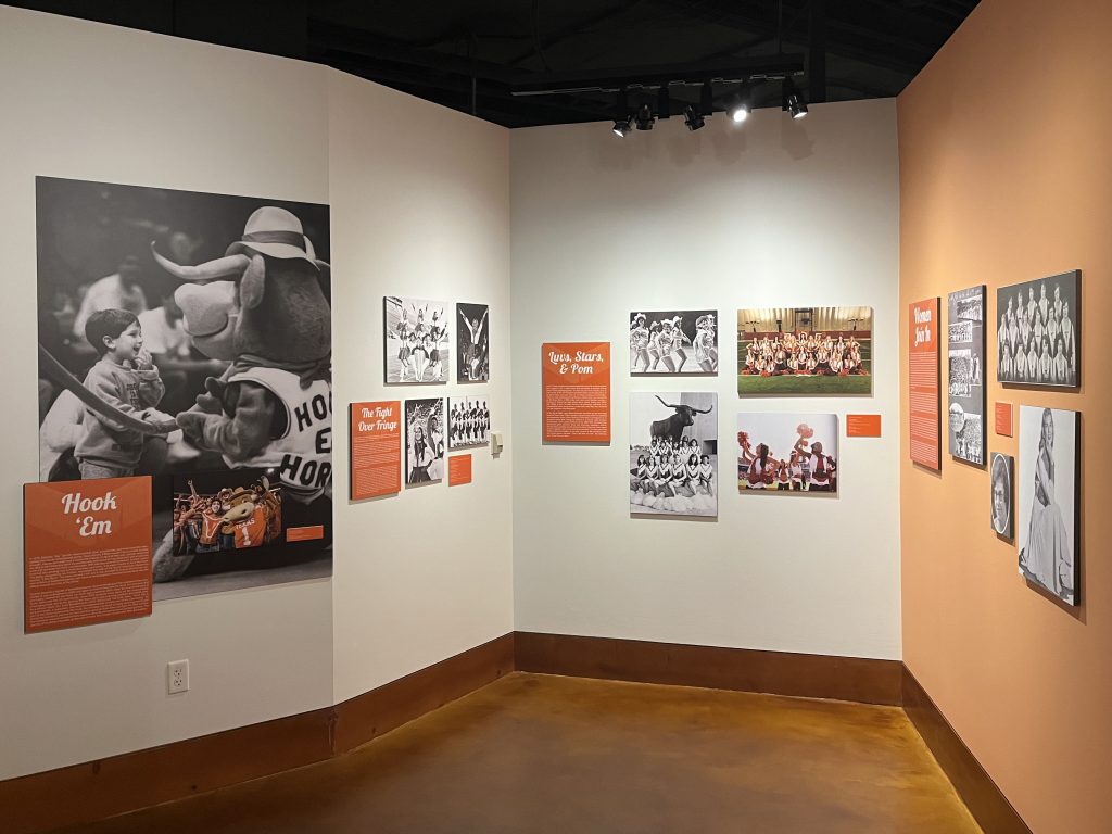 A color photo of several walls in the new exhibit, showing various black and white and color photograph reproductions with an especially large shot of the Hook Em mascot.