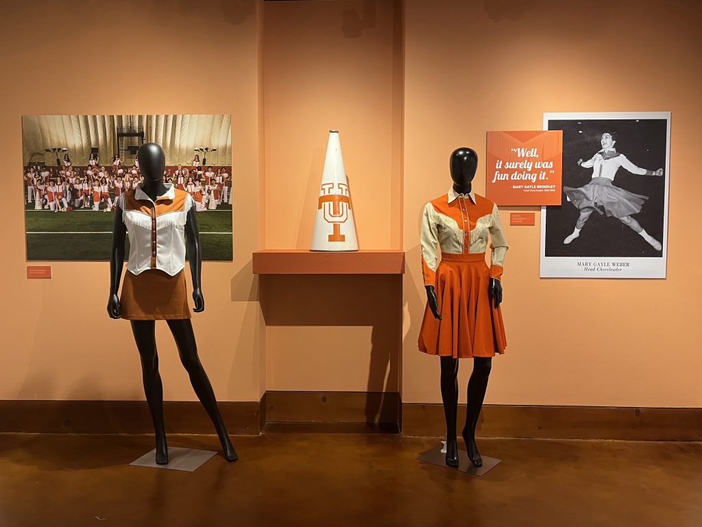 A soft orange wall with two female figures displaying cheer uniforms one from 1961 and the other from 2026. A megaphone on a floating shelf.